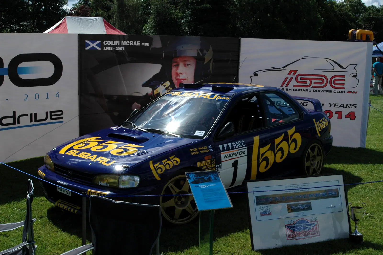 Colin McRae's blue and yellow 555 Subaru Impreza rally car on display at the ISDC club stand, with ISDC and Prodrive banners behind it